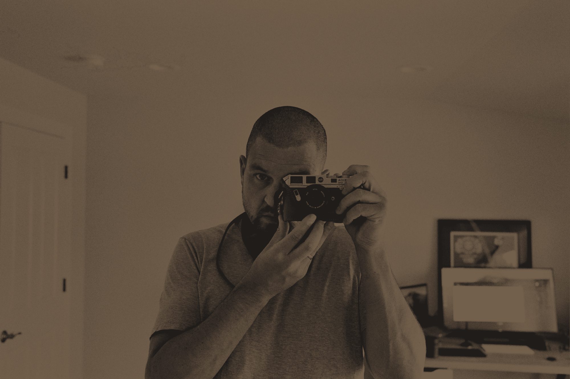 Mirror self-portrait of Matthew raising a Leica rangefinder to his eye, in black and white with a warm brown tint