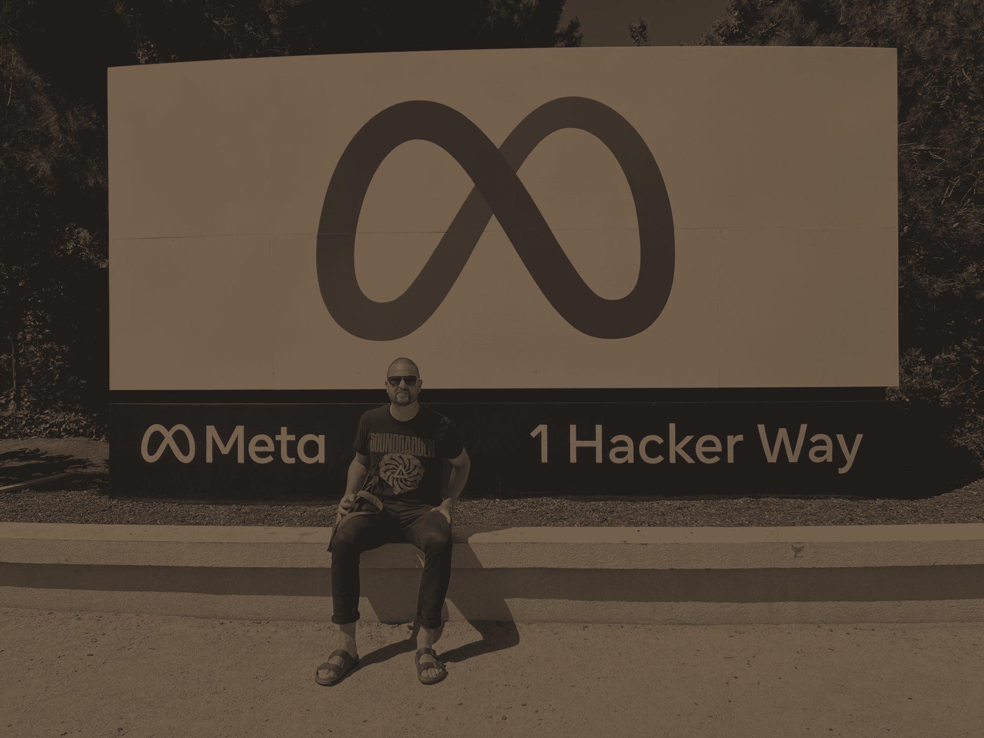 Matthew sitting in front of the Meta sign at 1 Hacker Way, in black and white with a warm brown tint